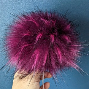 May include: A fluffy pom pom with a deep purple and black color combination. The pom pom is made of faux fur and has a soft, plush texture.