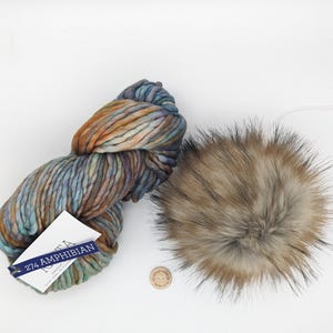 Puede incluir: Un ovillo de hilo multicolor en tonos azules, verdes, naranjas y marrones. El hilo está etiquetado como "274 AMPHIBIAN". También se muestra un pompón de piel sintética marrón y gris.