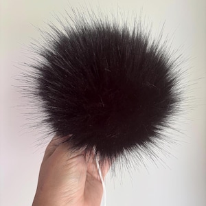 May include: A large, round, fluffy black pom-pom made of long, soft synthetic fibres. The spherical accessory features a dense, voluminous texture with a uniform dark black colour throughout, and it includes a thin white string attached at the base.
