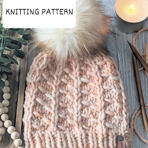 Könnte beinhalten: Eine gestrickte Mütze mit einem flauschigen Bommel in einem pfirsichfarbenen Rosa mit einem braunen Lederanhänger, auf dem "Cozy Knits" steht.