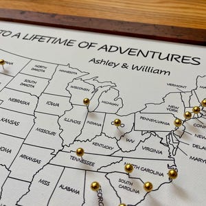 May include: A framed canvas map of the United States with gold push pins marking locations. The map is labeled "To a Lifetime of Adventures" and personalized with the names "Ashley & William".