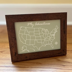 May include: A wooden frame with a light green map of the United States inside. The map is outlined in white and has the text "My Adventures" at the top.