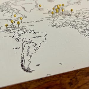 May include: A white world map with gold push pins marking locations. The map shows the continents of North America, South America, Europe, Africa, Asia, and Australia. The map is on a wooden surface.