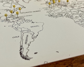 May include: A white world map with gold push pins marking locations. The map shows the continents of North America, South America, Europe, Africa, Asia, and Australia. The map is on a wooden surface.