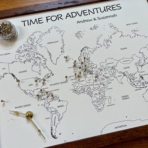 May include: A white world map with black outlines of countries and continents. The map is framed in a dark wood frame. The map has gold push pins placed on it, and the text "TIME FOR ADVENTURES" is at the top of the map. The text "Andrew & Susannah" is below the title.