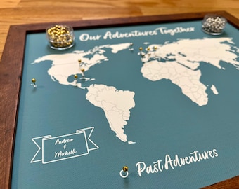 May include: A teal blue world map with white outlines of countries and continents. The map is framed in dark wood and has gold push pins marking locations. The text "Our Adventures Together" is at the top of the map and the text "Past Adventures" is at the bottom of the map. A banner with the text "Andrew & Michelle" is in the lower left corner of the map.