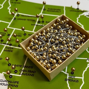 May include: A collection of gold-colored push pins in a small cardboard box, with some pins placed on a green map. The map features state names and geographical features.