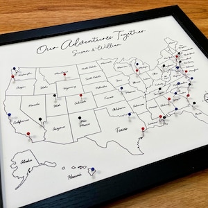 May include: A white map of the United States with black state borders and the text "Our Adventures Together Susan & William".  The map is framed in black and has red, blue, and black push pins marking locations.