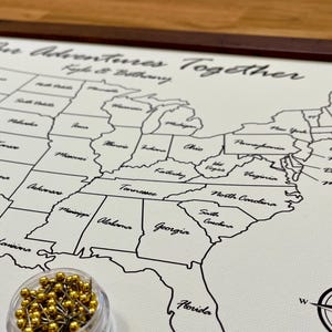 May include: A white map of the United States with black state outlines and state names. The map is framed in a dark wood frame. A small jar of gold push pins is in the lower left corner of the image. The map has the text "Our Adventures Together" at the top.