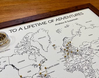 May include: A framed world map with a dark wood border, featuring black outlines of continents and countries. The map is marked with gold push pins. The text "TO A LIFETIME OF ADVENTURES" and names are printed at the top. A small jar of gold pins is in the corner.
