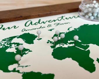 May include: A green world map with white push pins marking locations. The map is framed in a dark wood frame. The text "Our Adventure Amanda & Steven" is printed at the top of the map.