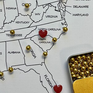 May include: A white map of the United States with state outlines and names. Gold push pins mark locations, with two red heart-shaped pins in North Carolina and Florida. A small gold tin holds additional gold pins.