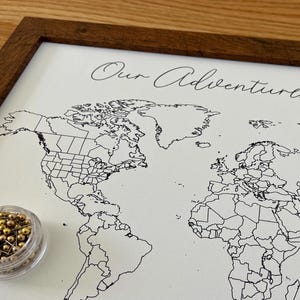 May include: A framed world map with the text "Our Adventures Together" in cursive. The map is black and white, with a dark brown wooden frame. A small clear container of gold push pins sits in the lower left corner.