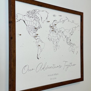 May include: A framed world map with push pins marking locations. The map is white with black lines and text. The frame is brown wood. The text below the map reads "Our Adventures Together Anna & William Est 2024".