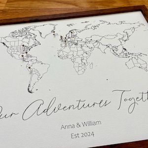 May include: A white framed map of the world with push pins marking locations. The text "Our Adventures Together" is written in cursive below the map, followed by "Anna & William Est 2024".