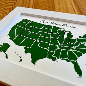 May include: A white framed map of the United States with the states outlined in white and filled in with dark green. The map includes Alaska and Hawaii. The text "Our Adventures" is written in cursive at the top of the map. A compass is in the bottom right corner.