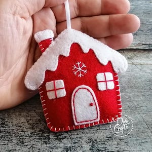 May include: A red and white felt house ornament featuring a snow-covered roof, a chimney, two square windows, and an arched door. The house is decorated with white stitched snowflake details and includes a white ribbon loop for hanging purposes.