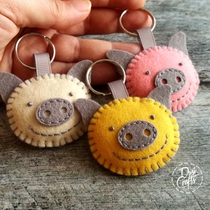 May include: Three felt pig keychains in cream, pink, and yellow. Each pig has gray ears and snout, with stitched details and a metal keyring. The keychains are handmade and feature a smiling face design.