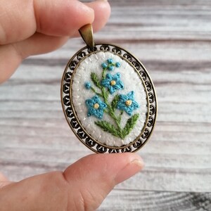 Peut inclure: Pendentif ovale avec un motif brodé de myosotis bleus sur fond blanc. Le pendentif est serti dans un cadre filigrané de couleur bronze, créant une esthétique vintage. Le pendentif est tenu par une main.