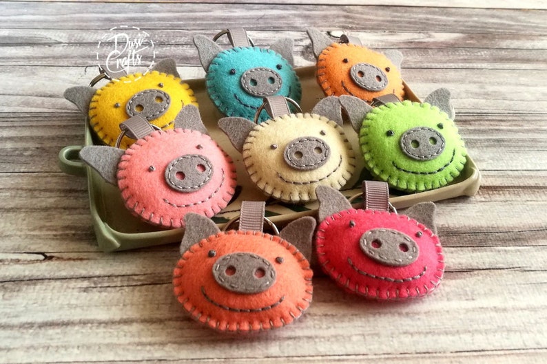 May include: A collection of colorful felt pig keychains in shades of yellow, blue, orange, pink, green, and red. Each keychain features a gray snout, ears, and a stitched smile. They are arranged in a green tray on a wooden surface.