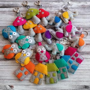 May include: A collection of felt keychains featuring various shapes including mushrooms, owls, elephants, and houses. Each item is crafted from grey felt with colorful accents in red, orange, yellow, green, blue, pink, and purple, attached to metal rings.
