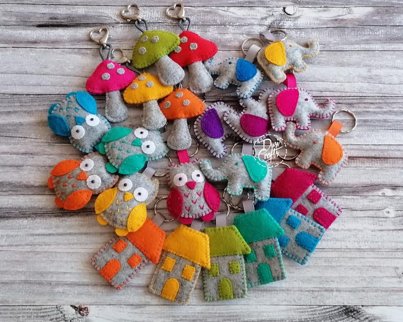 May include: A collection of felt keychains featuring various shapes including mushrooms, owls, elephants, and houses. Each item is crafted from grey felt with colorful accents in red, orange, yellow, green, blue, pink, and purple, attached to metal rings.