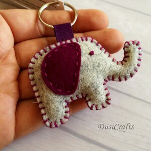 May include: A felt elephant keychain featuring a light gray body with a dark purple ear and matching purple blanket stitching along the edges. The elephant is attached to a silver metal split ring by a purple fabric ribbon loop.