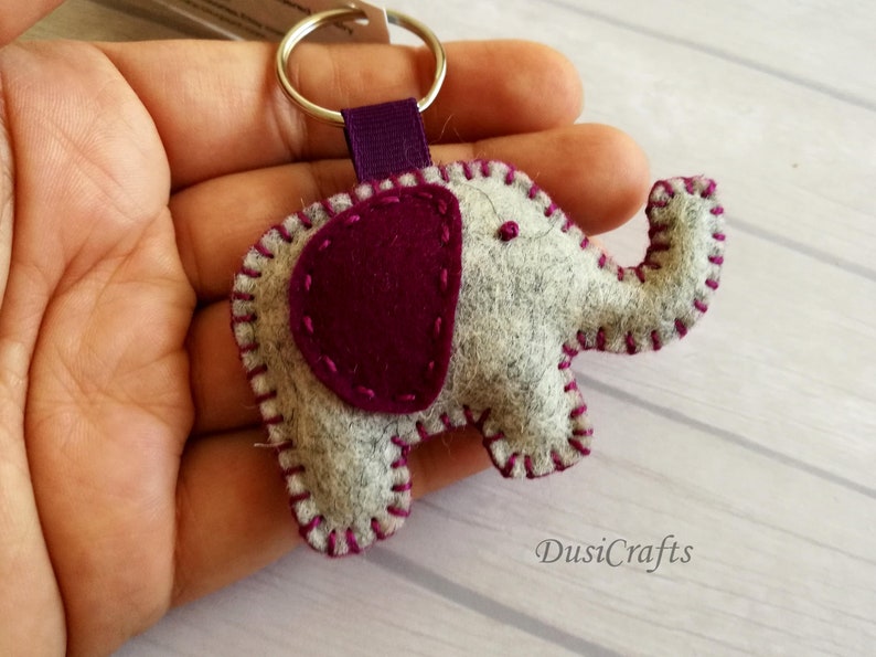 May include: A felt elephant keychain featuring a light gray body with a dark purple ear and matching purple blanket stitching along the edges. The elephant is attached to a silver metal split ring by a purple fabric ribbon loop.