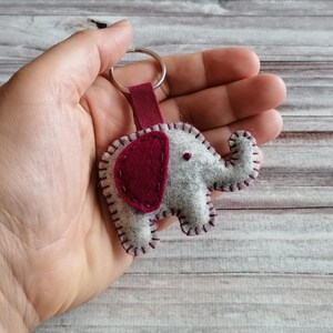 May include: A felt elephant keychain featuring a light gray body with a dark maroon ear and matching maroon whip-stitch detailing along the edges. The elephant is attached to a silver-toned metal split ring by a short, dark maroon fabric strap.
