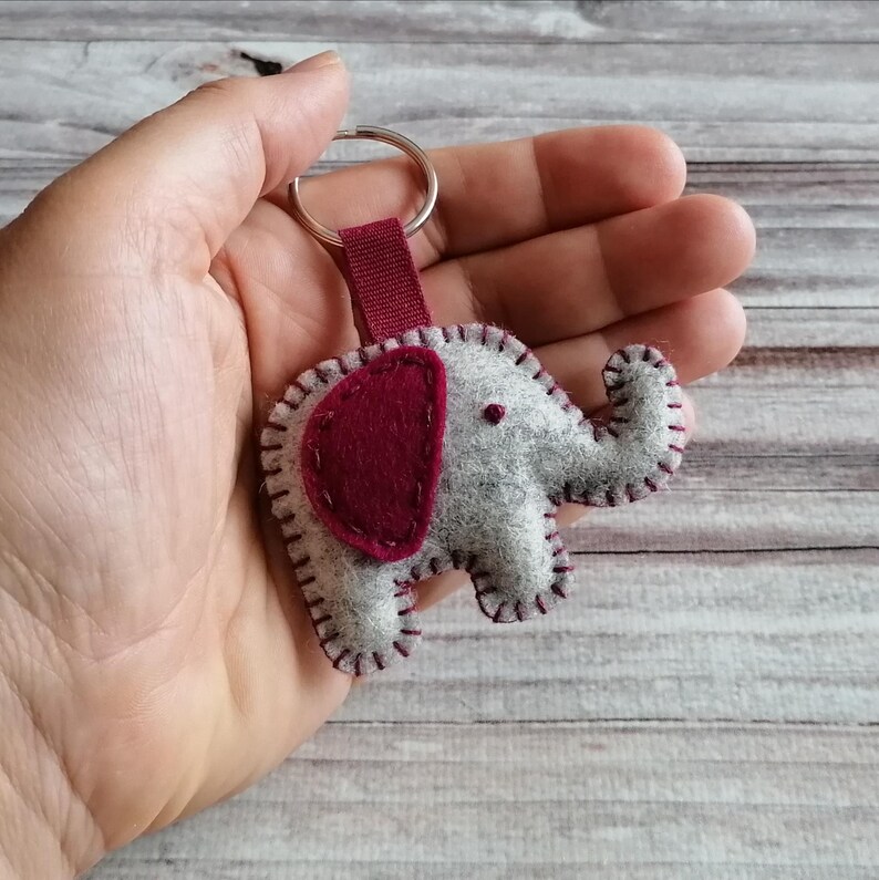 May include: A felt elephant keychain featuring a light gray body with a dark maroon ear and matching maroon whip-stitch detailing along the edges. The elephant is attached to a silver-toned metal split ring by a short, dark maroon fabric strap.