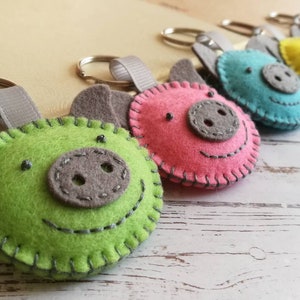 May include: A collection of colorful felt pig keychains. Each keychain features a pig face with stitched details, gray ears, and a gray snout. The pigs are in shades of green, pink, blue, yellow, and red. A gray ribbon loop and metal keyring are attached.