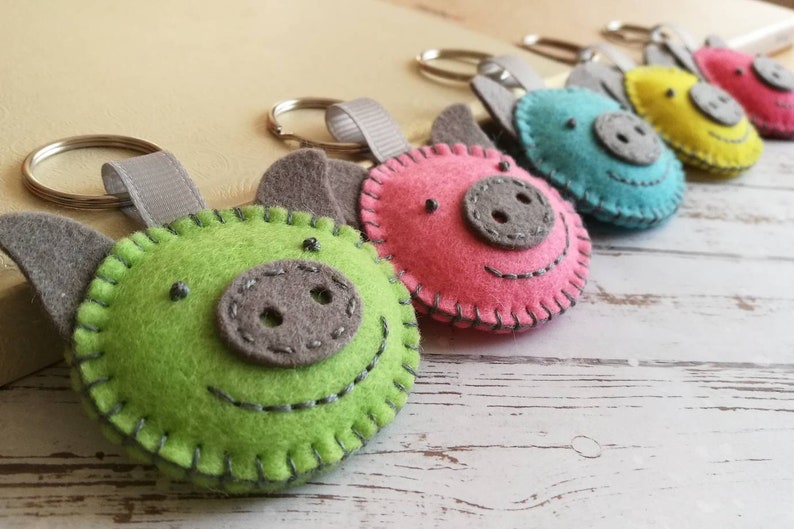 May include: A collection of colorful felt pig keychains. Each keychain features a pig face with stitched details, gray ears, and a gray snout. The pigs are in shades of green, pink, blue, yellow, and red. A gray ribbon loop and metal keyring are attached.
