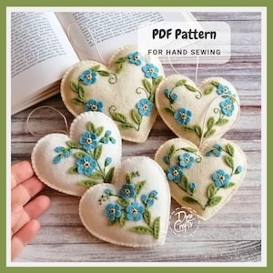 Puede incluir: Adornos de fieltro en forma de corazón de color crema con bordados florales azules y hojas verdes. Cada corazón tiene un lazo para colgar. La imagen incluye el texto "PDF Pattern For Hand Sewing".
