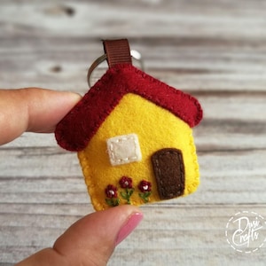 May include: A handmade felt house keychain. The house is yellow with a red roof, a white window, and a brown door. Three small red flowers are stitched below the door. A brown ribbon loop and silver keyring complete the design.