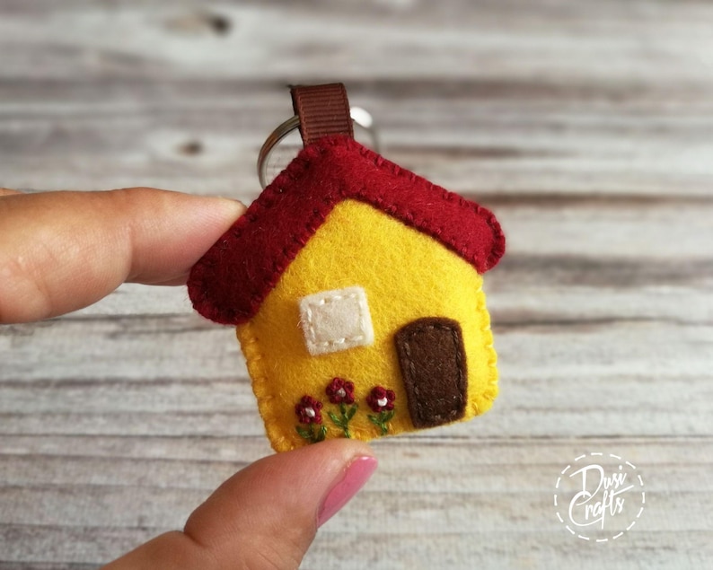 May include: A handmade felt house keychain. The house is yellow with a red roof, a white window, and a brown door. Three small red flowers are stitched below the door. A brown ribbon loop and silver keyring complete the design.