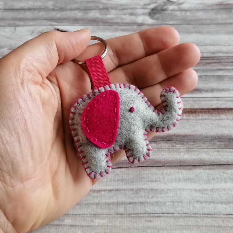 May include: A felt elephant keychain featuring a light gray body with a dark pink ear and matching dark pink blanket stitching along the edges. The elephant is attached to a silver metal split ring by a short, dark pink fabric strap.