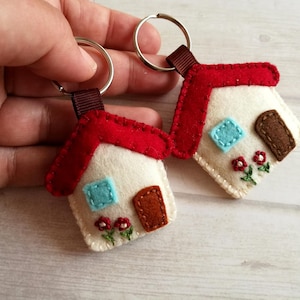 May include: Two felt house keychains. Each keychain features a white house with a red roof, a blue square window, and a brown door. The houses are decorated with embroidered flowers and attached to a metal keyring.