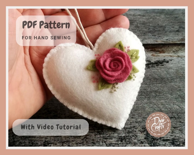 May include: White felt heart with a pink felt rose and green felt leaves. The heart is hand-sewn and has a string attached. The text "PDF Pattern for Hand Sewing" and "With Video Tutorial" is visible on the image.