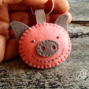 May include: A handmade felt pig keychain. The pig has a pink body, gray ears and snout, and a stitched smile. The keychain includes a gray ribbon loop and a silver keyring. The pig's face is hand-stitched with gray thread.