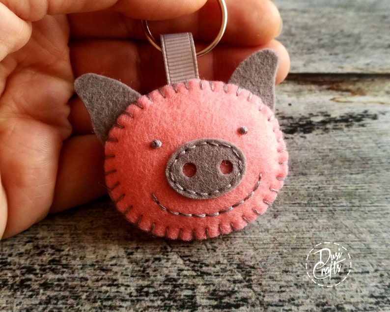 May include: A handmade felt pig keychain. The pig has a pink body, gray ears and snout, and a stitched smile. The keychain includes a gray ribbon loop and a silver keyring. The pig's face is hand-stitched with gray thread.