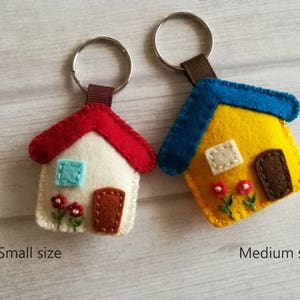 May include: Two felt house keychains. One is white with a red roof, blue window, and brown door. The other is yellow with a blue roof, white window, and brown door. Both have a brown strap and silver keyring. The text "Small size" and "Medium size" are below each keychain.