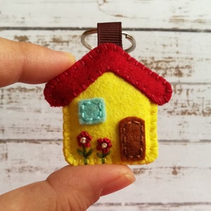May include: A handmade felt house keychain. The house is yellow with a red roof, a blue window, and a brown door. Two small red flowers with green stems are below the window. A brown loop and silver keyring are attached.