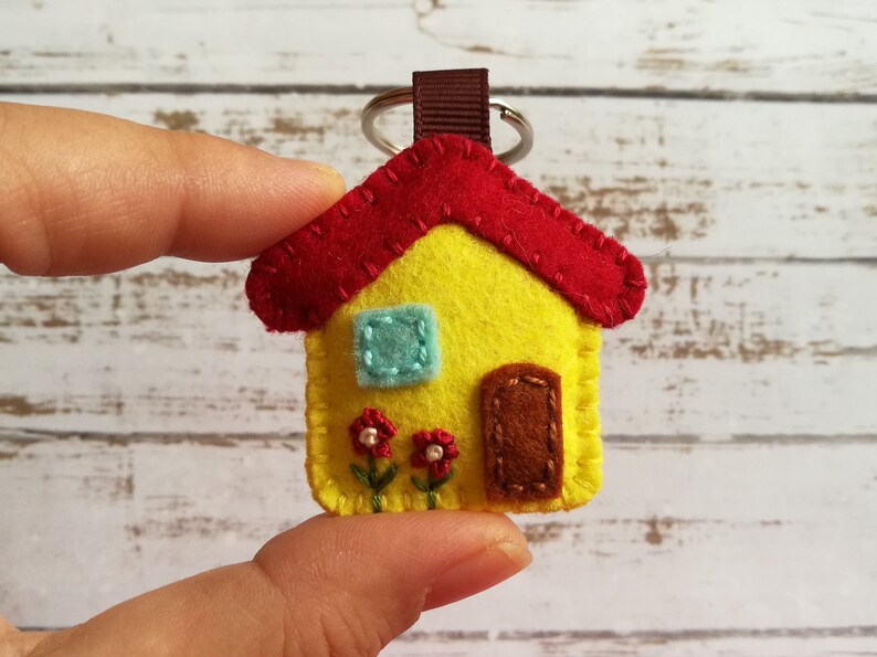 May include: A handmade felt house keychain. The house is yellow with a red roof, a blue window, and a brown door. Two small red flowers with green stems are below the window. A brown loop and silver keyring are attached.