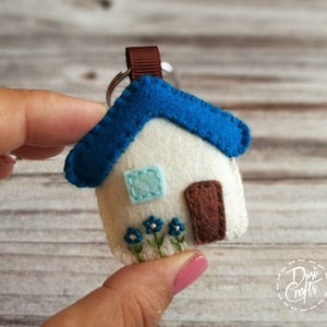 May include: A handmade felt house keychain. The house features a white body, a blue roof, a brown door, and a light blue window. Embroidered blue flowers adorn the bottom. A brown ribbon and silver keyring complete the design.