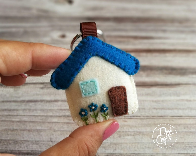 May include: A handmade felt house keychain. The house features a white body, a blue roof, a brown door, and a light blue window. Embroidered blue flowers adorn the bottom. A brown ribbon and silver keyring complete the design.