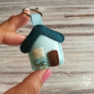 May include: A handmade felt house keychain in light blue with a teal roof and brown door. The house has a small white window and embroidered blue flowers. A light blue ribbon and silver keyring complete the design.