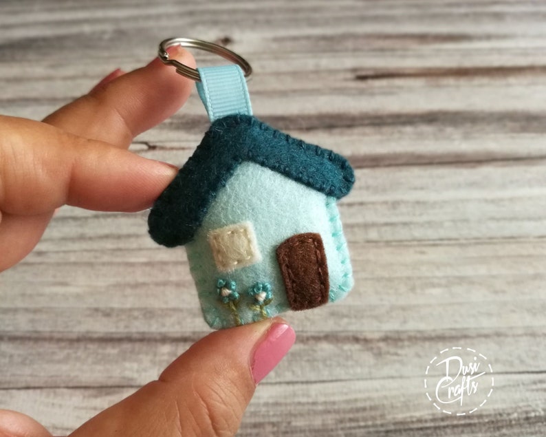 May include: A handmade felt house keychain in light blue with a teal roof and brown door. The house has a small white window and embroidered blue flowers. A light blue ribbon and silver keyring complete the design.