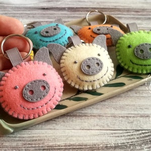 May include: A collection of handmade felt pig keychains in various colors: pink, blue, orange, green, and cream. Each keychain features a gray snout, ears, and a stitched smiling face. The keychains are displayed on a decorative tray.