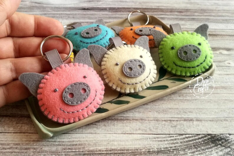May include: A collection of handmade felt pig keychains in various colors: pink, blue, orange, green, and cream. Each keychain features a gray snout, ears, and a stitched smiling face. The keychains are displayed on a decorative tray.