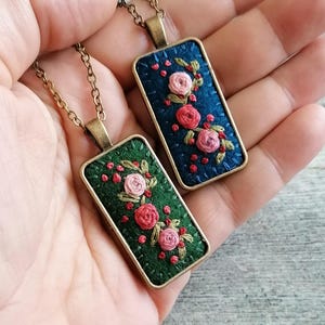 Op de afbeelding: Twee rechthoekige hangerkettingen met handgeborduurde bloemmotieven op stof. De ene hanger heeft een donkergroene achtergrond en de andere een donkerblauwe achtergrond, beide gevat in antiek bronzen frames met bijpassende schakelkettingen.
